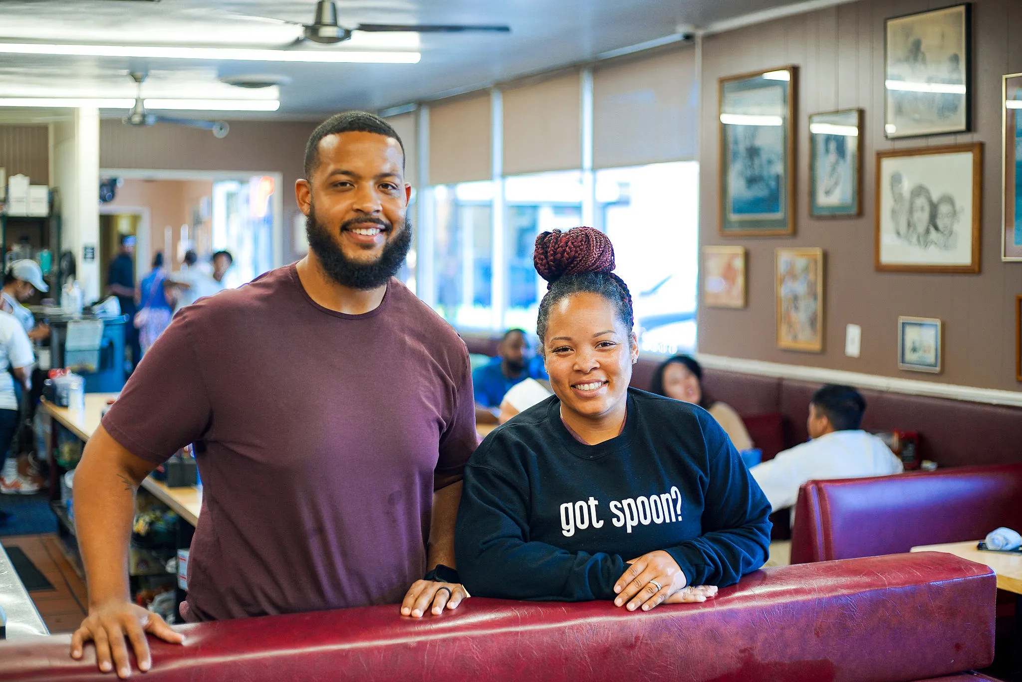 A smiling man and woman leaning on a red booth inside The Serving Spoon, she in a “got spoon?” sweatshirt